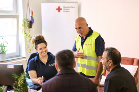 Eine Frau und ein Mann in Rotkreuzkleidung sitzen in einem Beratungsraum zwei Männern gegenüber und lächeln ihnen zugewandt zu.