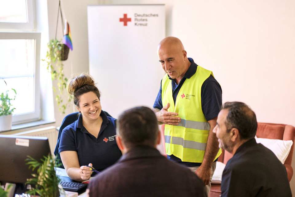 Eine Frau und ein Mann in Rotkreuzkleidung sitzen in einem Beratungsraum zwei Männern gegenüber und lächeln ihnen zugewandt zu.
