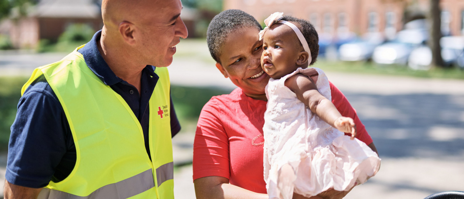 DRK-Mitarbeiter mit gelber Weste begrüßt ein Frau mit Baby vor einer Unterkunft